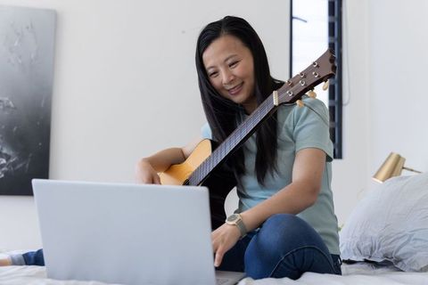 Mature asian woman practicing acoustic guitar during online lesson on bed with laptop