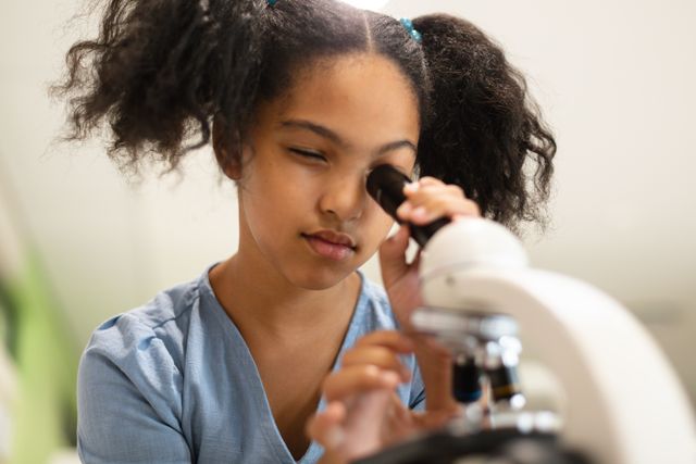Young biracial girl focusing on microscope in chemistry class, ideal for educational materials, STEM programs, school brochures, and science-related content. Highlights importance of early scientific education and curiosity in young students.