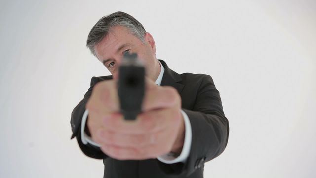 A businessman in a suit pointing a gun directly at the camera, creating a sense of threat and danger. Suitable for use in media portraying crime, action sequences, self-defense, or dramatic scenarios in movies or graphic content.