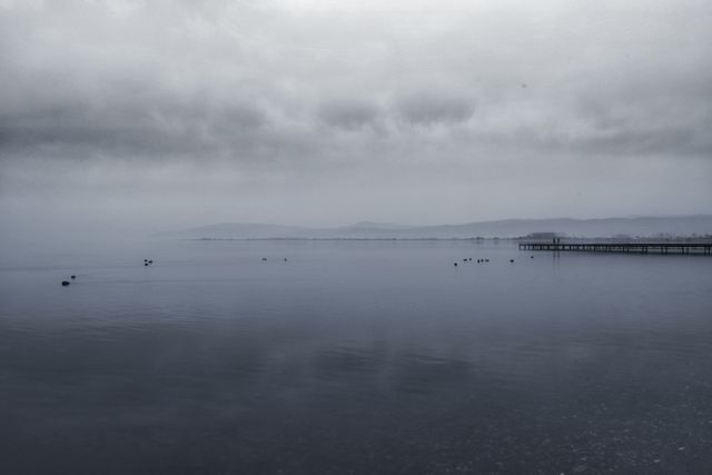 This image shows a calm lake with a misty, foggy atmosphere in the early morning. Mountains are visible in the background, and the scene exudes tranquility and serenity. Perfect for use in calming or meditative content, nature articles, or backgrounds which need a peaceful and atmospheric appeal.
