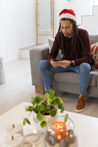 Mid-adult man wearing santa hat checking smartphone on sofa in cozy modern living room