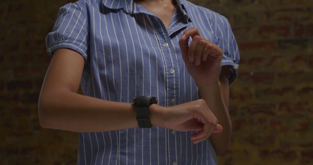 Close-up of a person checking the time on their smartwatch indoors. The individual is wearing a blue-striped shirt and is focusing on the watch. Useful for illustrating concepts of technology, time management, wearables, and modern lifestyle.