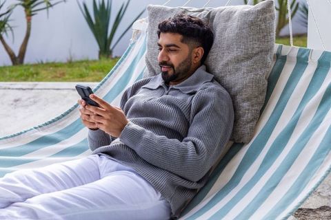 Man reclining on striped hammock using smartphone wearing gray pullover outdoors relaxing