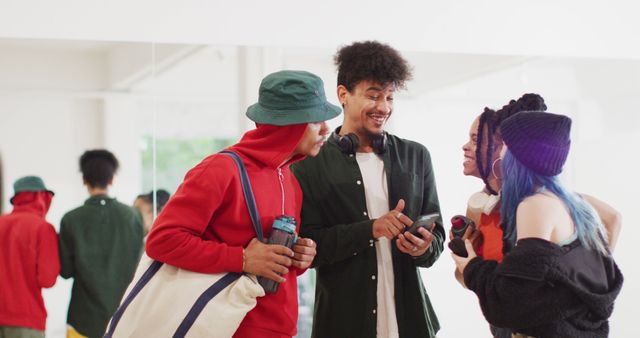 Group of young, diverse friends gathered in a dance studio, huddled around a smartphone enjoying each other's company. They wear casual, trendy clothing that includes bucket hats and vibrant colors. Ideal for promoting friendship, social bonding, and youth culture. Can be used in advertisements for fashion brands, social apps, or community events.