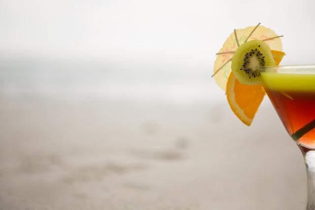 Glass of cocktail drink garnished with kiwi, lime and cocktail umbrella at tropical beach