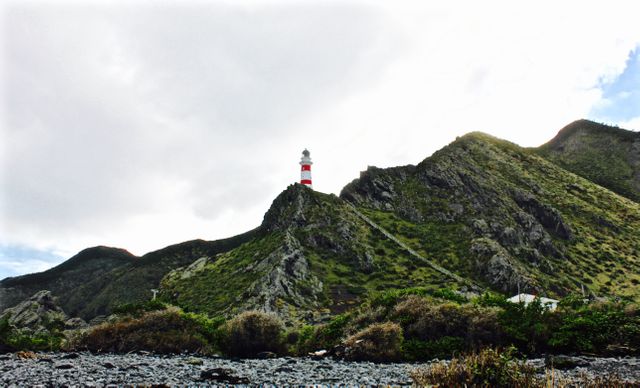 Lighthouse stands tall on the peak of a rocky mountain against a backdrop of a cloudy sky. Lush greenery surrounds the frame, creating a contrast with the rough terrain. Use this for themes related to navigation, maritime safety, coastal landscapes, travel destinations, or nature photography.