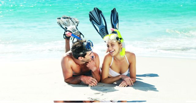 Couple lying on sandy beach wearing snorkeling gear, smiling at each other. Image ideal for travel websites, beach resort promotions, vacation planning materials, and advertisements focused on summer fun and relaxation.