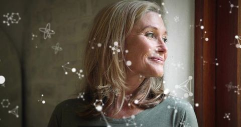 Mature woman gazing out sunlit window with digital molecular overlay suggesting reflection