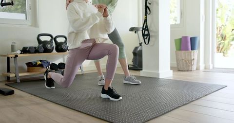 Home gym trainer guiding client in lunge on textured mat with kettlebells and trx