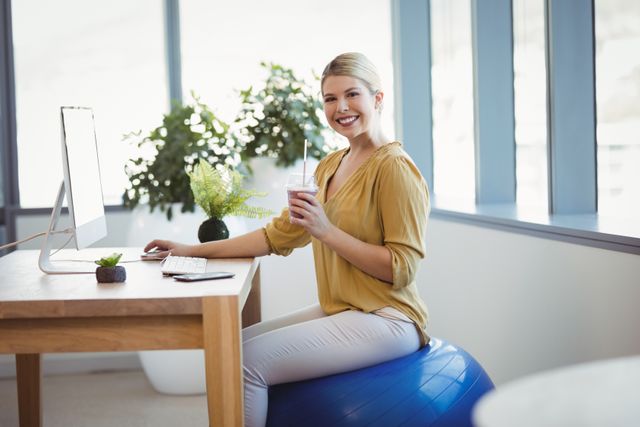 Businesswoman sitting on fitness ball at desk in modern office, promoting healthy lifestyle and ergonomic work environment. Ideal for articles on workplace wellness, productivity tips, and modern office design.