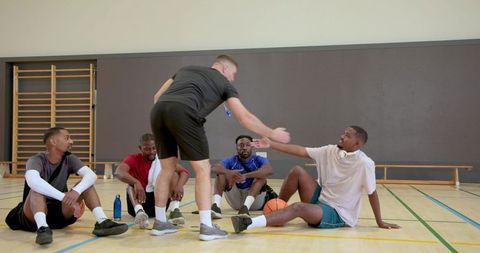Basketball coach offering hand to seated player in gym team huddle with teammates