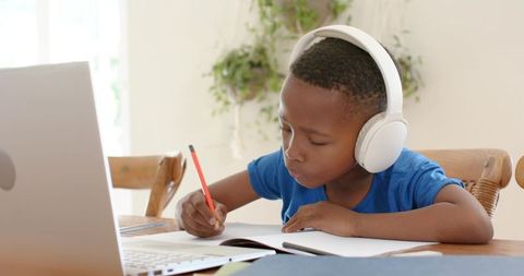 African american boy concentrating on homework wearing headphones while using laptop
