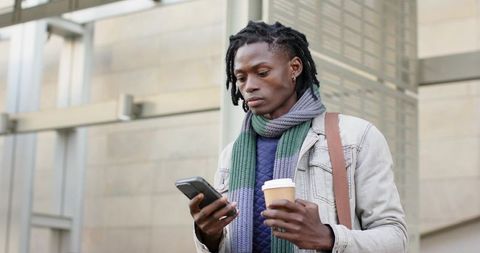 African american commuter checking smartphone while holding coffee cup at urban plaza