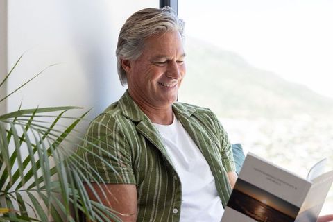 Smiling senior man reading paperback on sofa by bright window with indoor plants