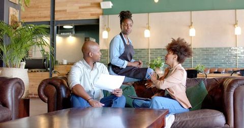 African American Barista Listening While Customers Discussing Documents on Cozy Coffeehouse Sofa