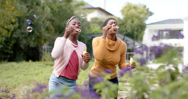 Happy african american daughter and mother blowing bubbles in sunny garden. Family, fatherhood, childhood, spring, happiness, lifestyle, relaxation, unaltered.