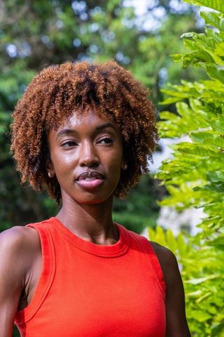 Sunlit african american woman posing in red sleeveless top among lush green foliage