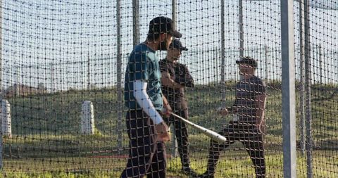 Three baseball players practicing batting in outdoor cage during sunny training session