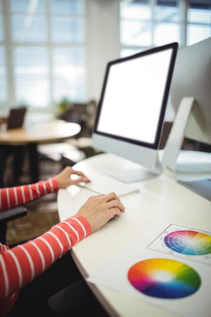Graphic designer is working on a computer in a well-lit office. A color wheel is visible on her desk, indicating she is engaged in creative tasks. Suitable for illustrating modern workspace, creativity in business environments, and professional design work.