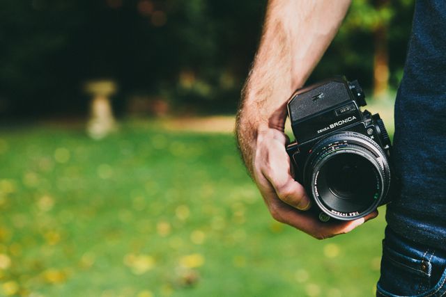 The photo depicts a photographer's hand holding a vintage camera, standing in a park filled with greenery. Ideal for use in articles related to photography as a hobby, vintage cameras, outdoor activities, manual photography techniques, or product advertisements for camera enthusiasts.