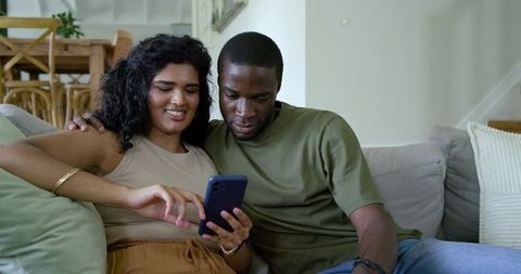Couple relaxing on sofa sharing smartphone, indian woman tapping screen, partner leaning in