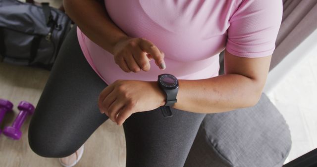 Woman wearing pink top and dark leggings is checking her smartwatch during a home workout. Various exercise accessories, including dumbbells and a gym bag visible nearby. Ideal for content related to fitness, health tracking, modern technology in sports, and home exercise routines.