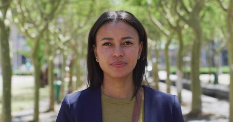 Confident young woman standing in city park wearing navy blazer and olive top, headshot