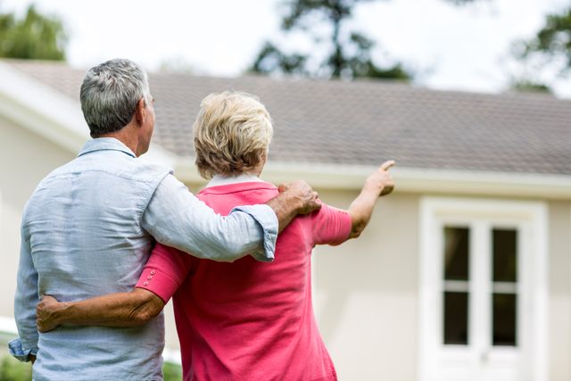 Senior couple standing in yard, looking at their new home with arms around each other. Ideal for real estate advertisements, retirement planning brochures, family lifestyle articles, and senior living promotions.
