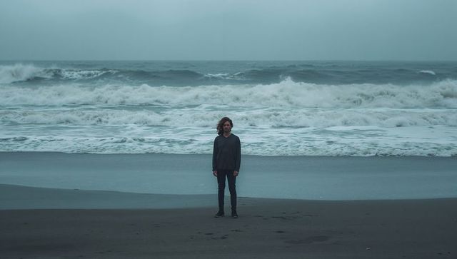 Solitary man standing on moody dark beach facing camera with crashing waves and overcast sky