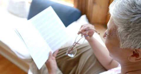 Senior Woman Relaxing Indoors Reading Book with Glasses in Hand