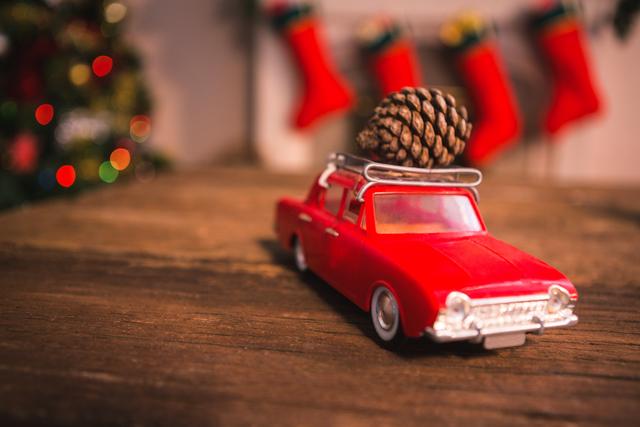 Toy car with a pine cone on its roof sitting on wooden table during Christmas time. Christmas stockings and bokeh lights in background add to festive atmosphere. Ideal for holiday greeting cards, Christmas decorations, festive advertisements, and winter-themed promotions.