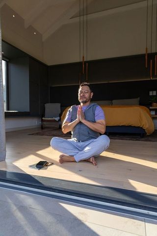 Asian man meditating in sunlit minimalist bedroom sitting cross-legged with smartphone and tablet