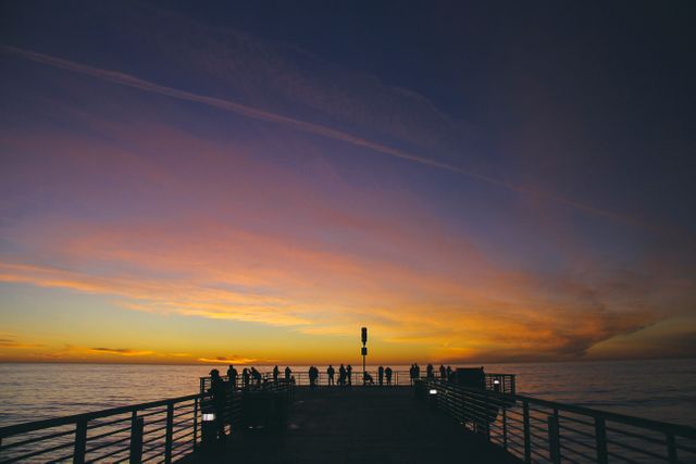Silhouetted people standing on pier watching vibrant sunset over the ocean. Fiery sky with calm waters makes it perfect for commercial use in travel brochures, website banners, backgrounds, or social media posts aimed at promoting tranquility and relaxation associated with coastal locations.