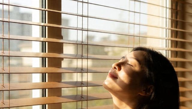 Basking asian woman tilting face toward sunlight through wooden blinds, serene portrait