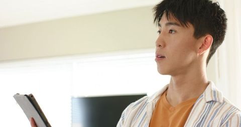 Young asian man holding tablet in bright living room wearing striped overshirt and orange shirt