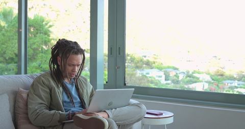 Man in 30s with dreadlocks working remotely on sofa by large window in minimalist interior