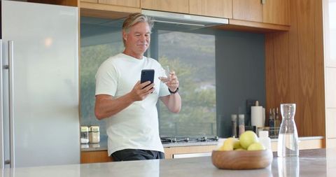 Senior man standing in modern kitchen checking smartphone while holding glass of water