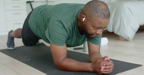 African american man holding forearm plank during home workout with earbuds and phone
