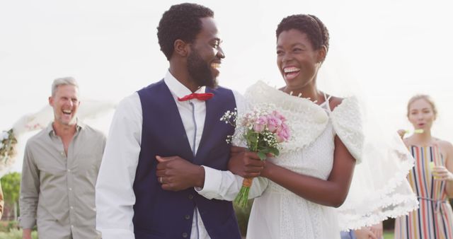 African American bride and groom smiling while walking outdoors dressed in wedding attire and holding a bouquet, with their friends celebrating in the background. Ideal for use in wedding websites, promotional materials, social media content related to weddings, and lifestyle blogs.