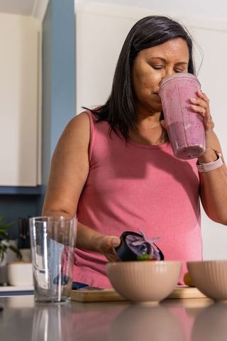 Mature woman tasting berry smoothie from blender cup at home kitchen counter in pink top
