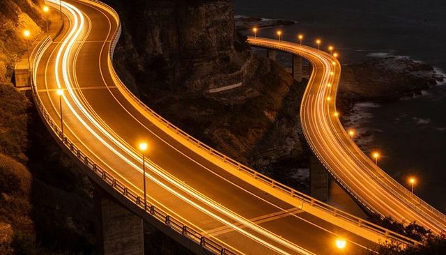 Winding coastal overpass at night with golden light trails and illuminated cliffside curve