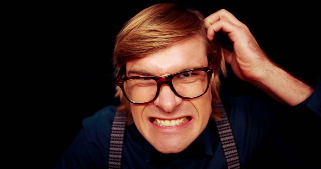 Man with blonde hair scratching head and grimacing while wearing large glasses and suspenders against dark background. Ideal for depicting confusion, frustration, problem-solving, brainstorming, emotions, stress. Useful for articles, advertisements, presentations related to tech support, troubleshooting, difficult decisions.