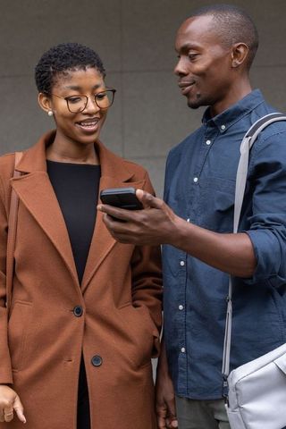 Two friends sharing smartphone and smiling while standing by concrete wall in city