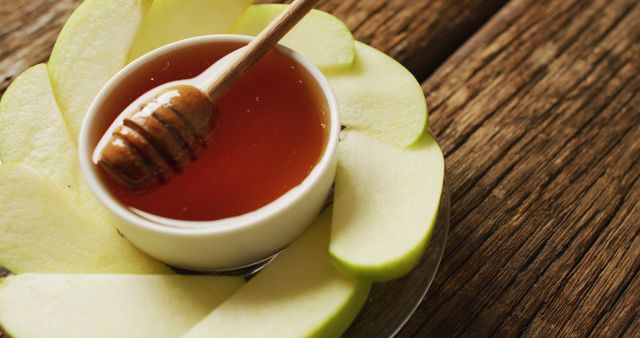 Close-up view of fresh apple slices arranged around a small bowl of honey on rustic wooden table. Honey dipper resting on bowl covered in honey. Suitable for displays related to healthy eating, organic food, natural ingredients, or seasonal treats.