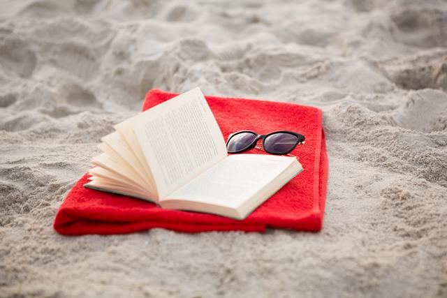 Open book with sunglasses on red towel at sandy beach. Perfect for concepts of relaxation, vacation, leisure time, summer holiday, outdoor reading, and serene getaway. Ideal for travel blogs, vacation planning websites, and summer promotions.