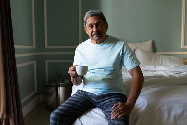 Senior man sitting on bed holding coffee cup, wearing casual pajamas. Ideal for content on retirement lifestyle, self-isolation during the pandemic, morning routines, and elderly well-being. Suitable for articles, blogs, and advertisements focusing on senior health, home comfort, and peaceful living.