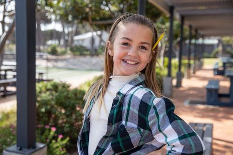 Cheerful school girl standing outdoors in courtyard with pencil