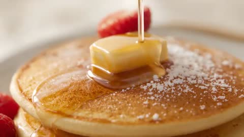 Pouring maple syrup over melting butter on stack of golden pancakes with raspberries