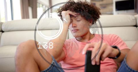 Young man sitting on couch rubbing forehead holding remote with futuristic hud overlay
