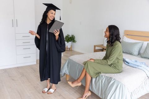 Diverse sisters celebrating graduation at home wearing cap and gown holding tablet smiling